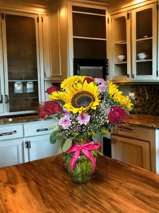 Arreglo mediano en base de vidrio con girasoles, rosas, gerberas, clavel, solidago y follaje verde.